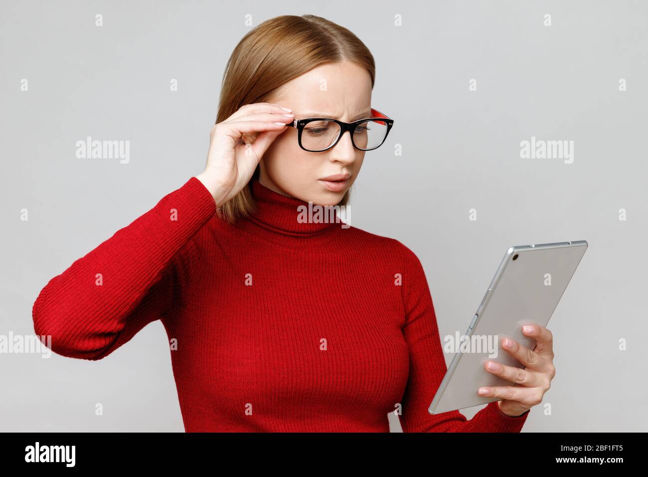 Stupito e sorpreso elegante donna in rosso tartareneck utilizzando il computer tablet, legge qualcosa di importante su coronavirus e l'economia, ho Foto Stock