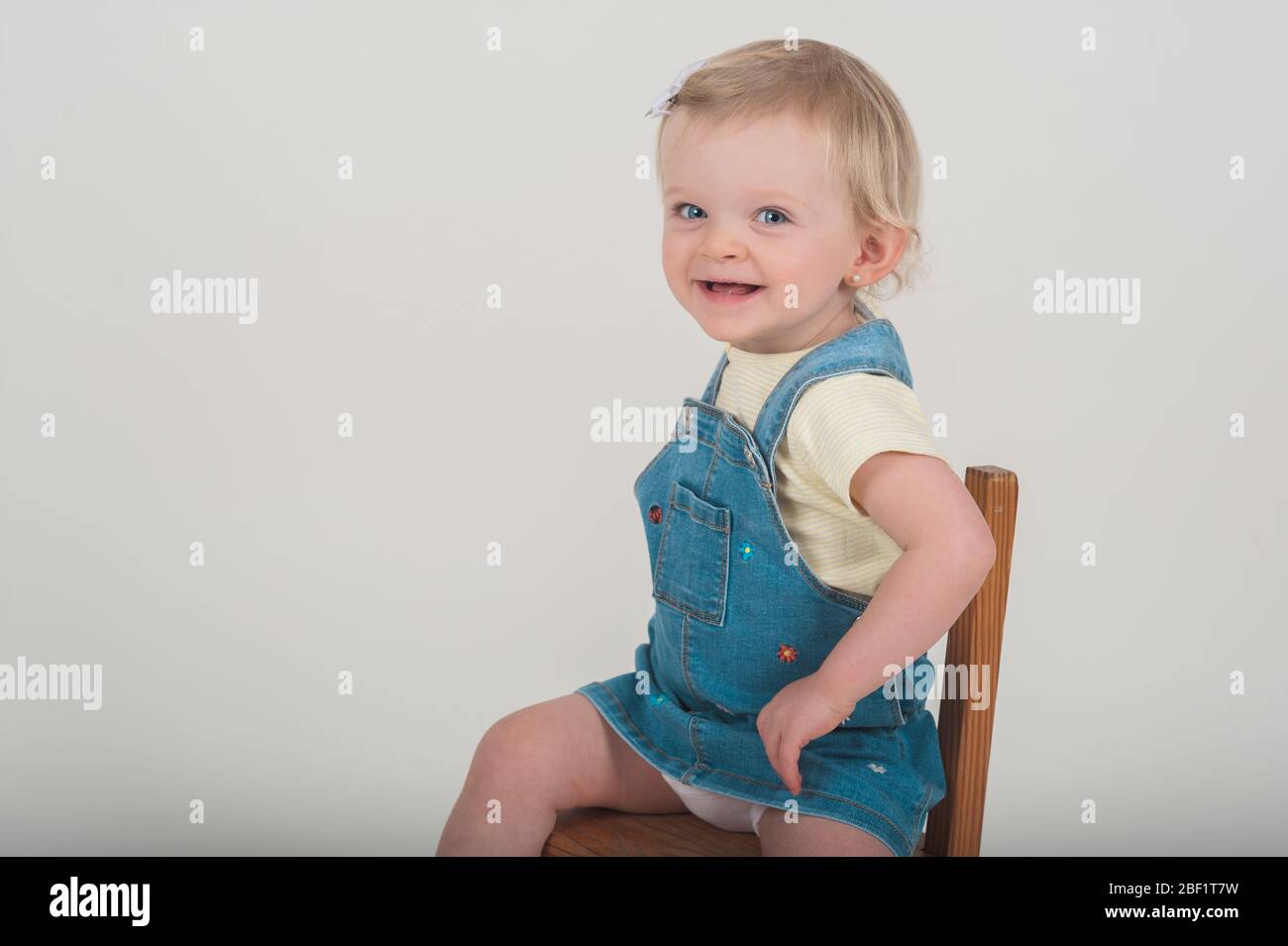 Adorabile bambina di 18 mesi sorridente alla sua mamma mentre si siede sulla sua sedia di legno nello studio di fotografia Aurelia Dumont a Playa del Rey, CA. Foto Stock