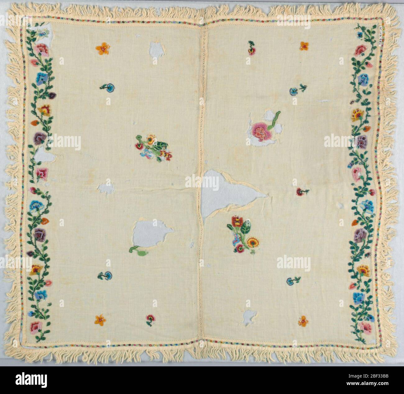 Dily. Tessuto quadrato di cotone bianco composto da due pezzi cuciti insieme con punti a pizzo. Fringed su tutti e quattro i lati. Fiori sparsi su terra. Due estremità rifilate con disegno di vite fiorita. Perline policrome utilizzate. Foto Stock