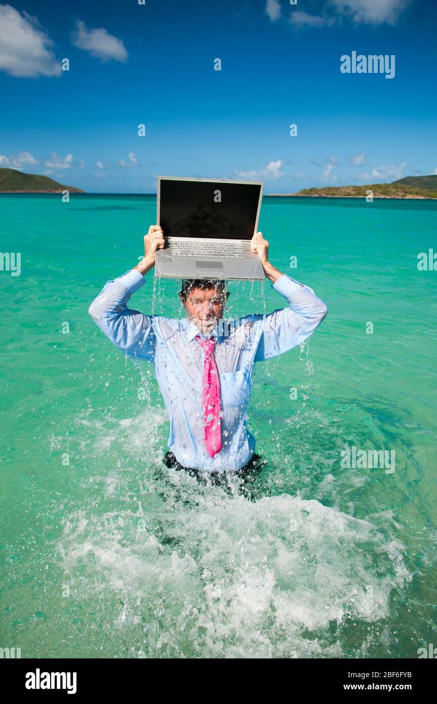 Uomo d'affari che schegge fuori dall'acqua con un portatile bilanciato sulla testa in mare tropicale blu brillante Foto Stock