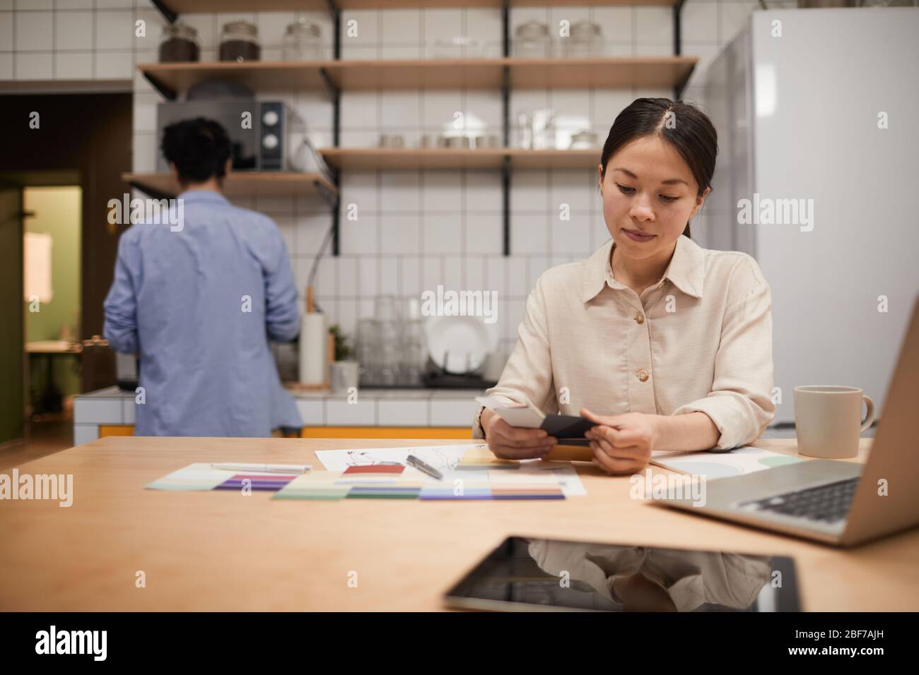 Giovane e seria donna d'affari seduta al tavolo con il laptop e lavorando con motivi colorati in cucina in ufficio Foto Stock