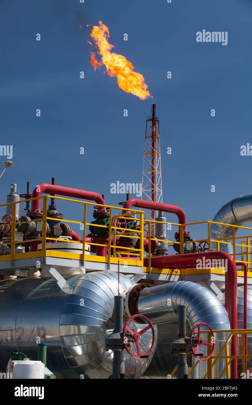 Stabilimento petrolchimico (raffineria petrolifera). Tubi, tubi, valvole e scambiatore di calore su torcia a gas gigante arancione e torre su sfondo cielo blu. Foto Stock