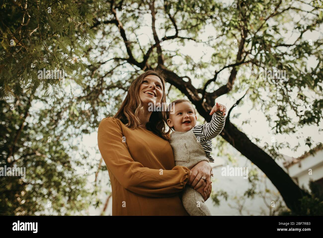 Madre che tiene il suo figlio del bambino in armi e bambino che ride, con albero verde sullo sfondo. Foto Stock