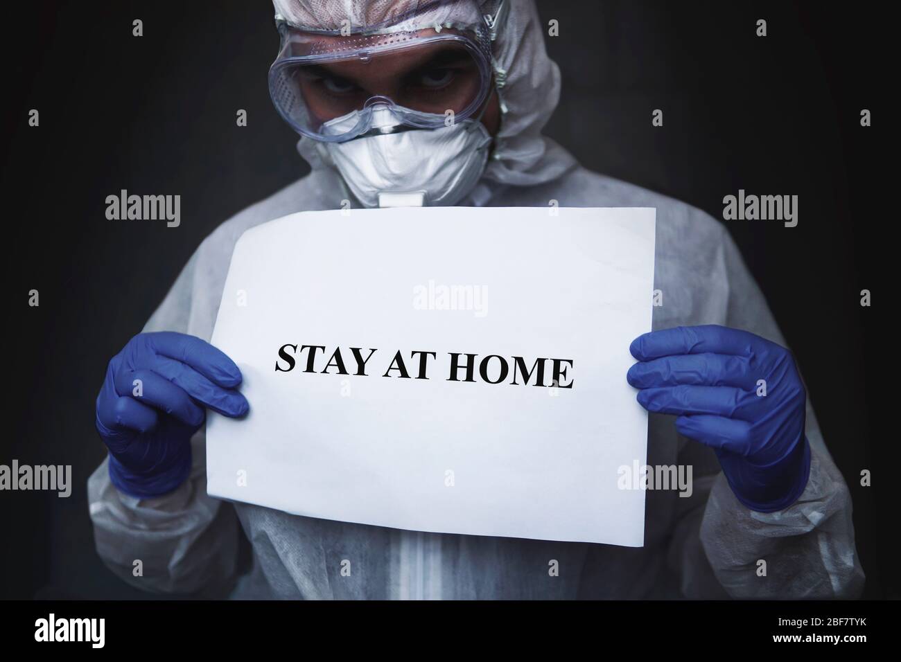 Un medico in una tuta biochimica protettiva e maschera con una carta con iscrizione stare a casa tenendo a portata di mano su uno sfondo nero scuro. Proteggere da Foto Stock