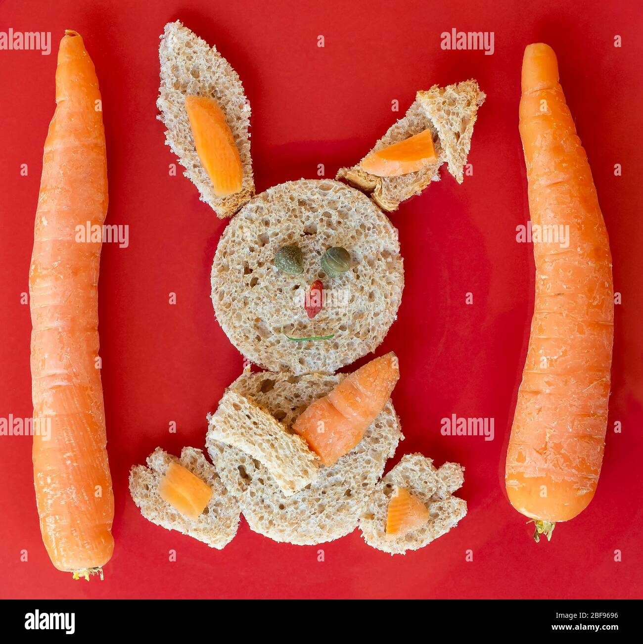Bel coniglietto fatto con pane panino integrale, capperi, carote e goji su sfondo rosso Foto Stock
