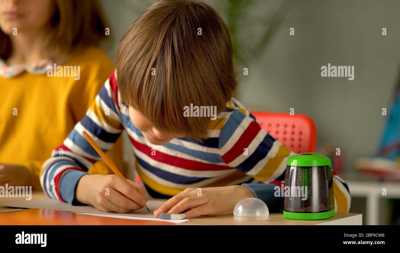 Apprendimento a distanza per gli scolari durante il periodo di quarantena del coronavirus. I bambini sono seduti al tavolo. In primo piano il ragazzo prende appunti a matita. Foto Stock