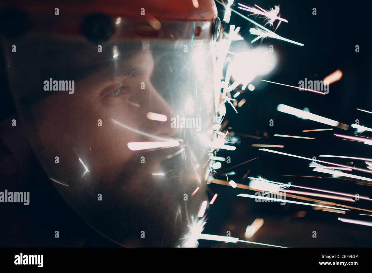 Uomo adulto bearded in maschera protettiva trasparente con particelle di metallo volanti scintille nell'oscurità Foto Stock