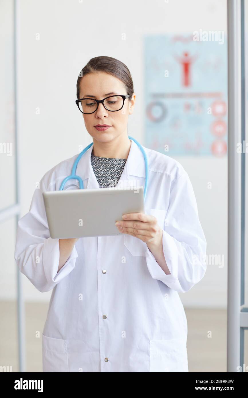 Medico donna serio in occhiali che indossano cappotto bianco e che lavora online su tablet digitale mentre si trova in ufficio Foto Stock