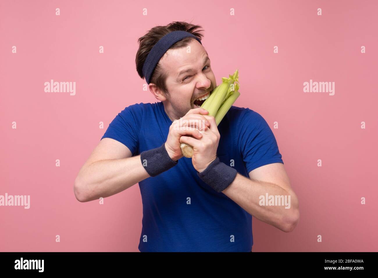 Giovane sportivo caucasico con sedano su fondo rosa. Amo tanto le verdure. Concetto di dieta verde. Foto Stock