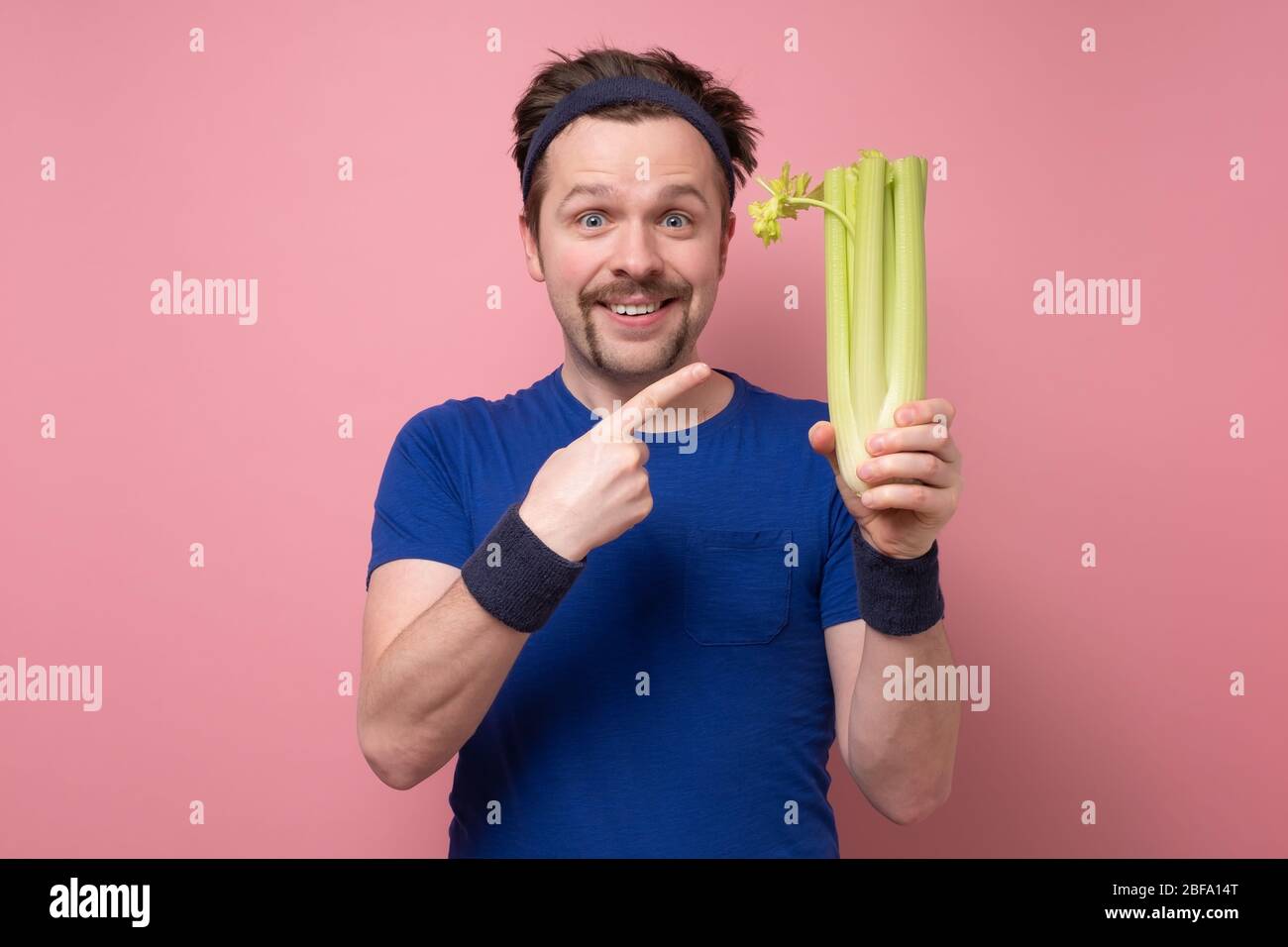 Giovane sportivo caucasico con sedano su fondo rosa. Amo tanto le verdure. Concetto di dieta verde. Foto Stock