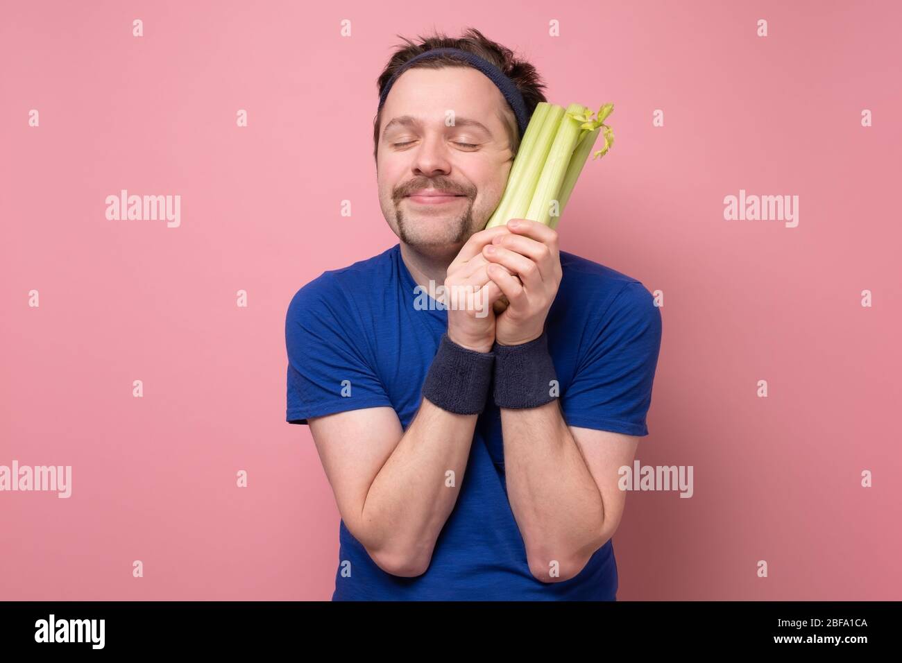 Giovane sportivo caucasico con sedano su fondo rosa. Amo tanto le verdure. Concetto di dieta verde. Foto Stock