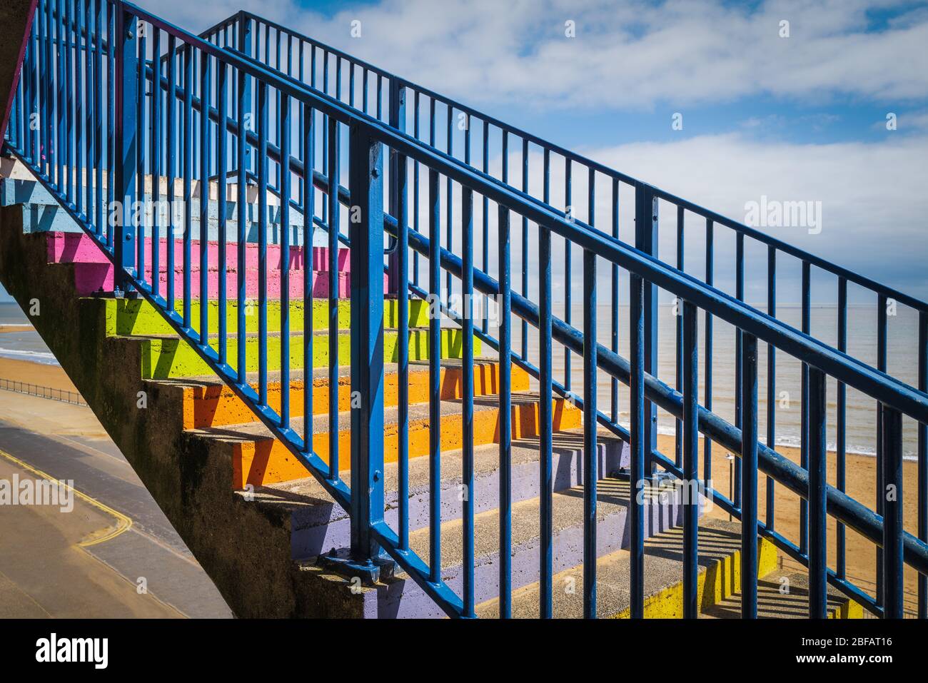 Gradini in cemento dipinti colori arcobaleno con una ringhiera blu davanti ad una spiaggia di sabbia, il cielo è blu con nuvole bianche. Foto Stock