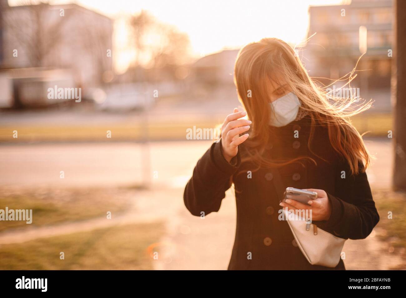 Giovane donna che indossa una maschera protettiva per il viso mentre usa lo smartphone mentre cammina per strada in città Foto Stock