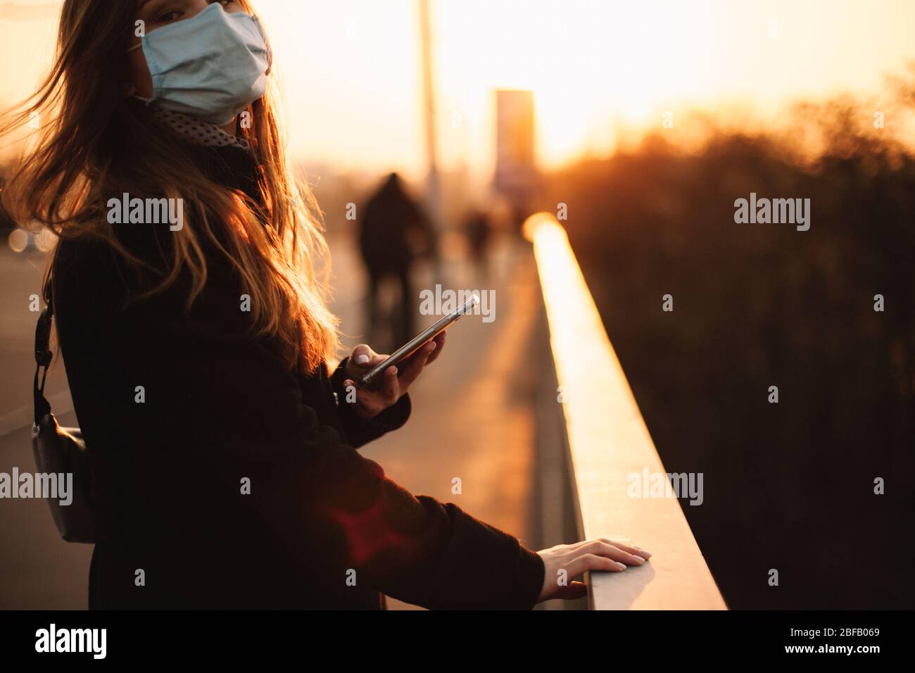Giovane donna che indossa la maschera medica protettiva del viso con lo smartphone mentre tiene la ringhiera in piedi sul ponte in città al tramonto Foto Stock