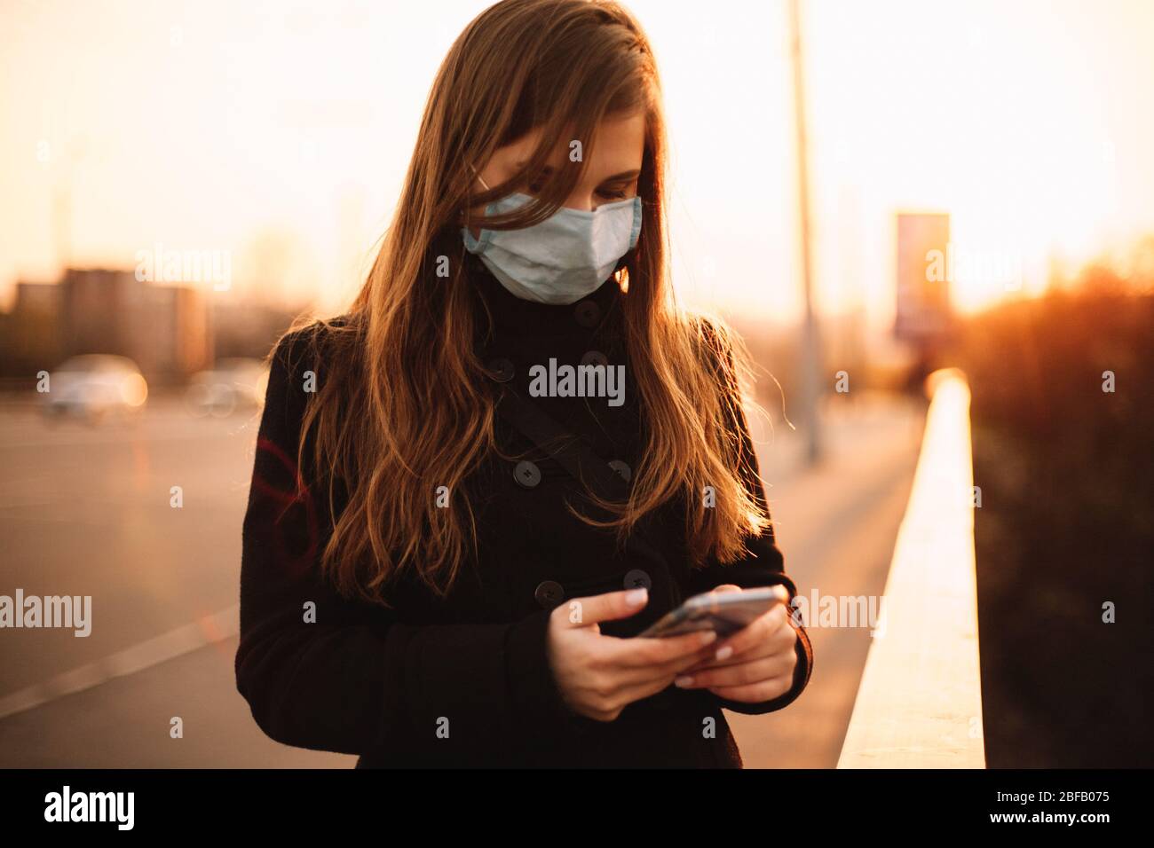 Adolescente ragazza indossare maschera protettiva viso medico utilizzando smartphone mentre in piedi sul ponte in città al tramonto Foto Stock