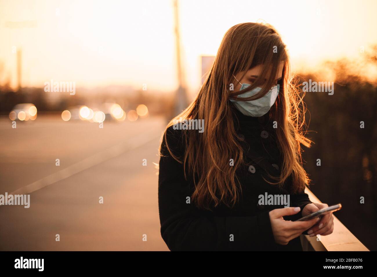 Adolescente ragazza indossare maschera protettiva viso medico utilizzando smartphone mentre in piedi sul ponte in città al tramonto Foto Stock
