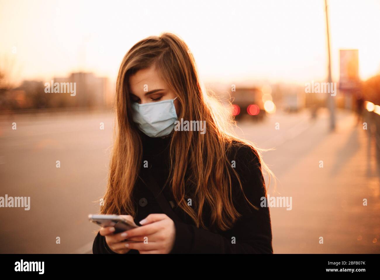 Giovane donna seria che indossa la maschera protettiva facciale medica utilizzando lo smartphone mentre cammina sul marciapiede vuoto in città al tramonto Foto Stock
