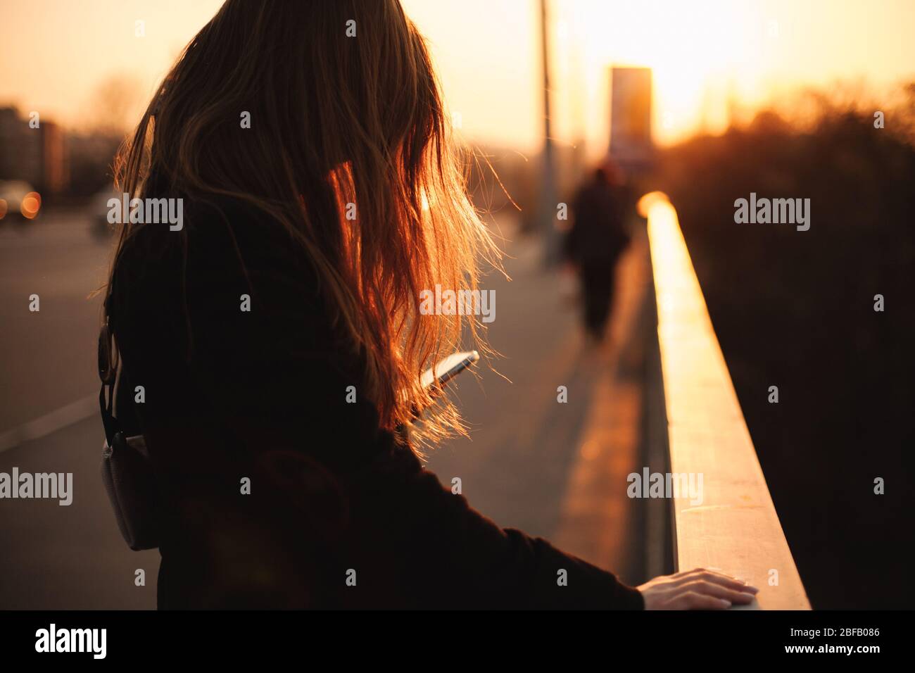 Giovane donna che usa lo smartphone mentre tiene la ringhiera in piedi sul ponte in città al tramonto Foto Stock