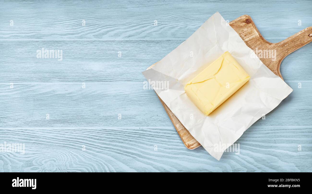 Pezzo di burro su carta da imballaggio e asse di legno. Vista dall'alto del burro su sfondo di legno blu. Foto Stock