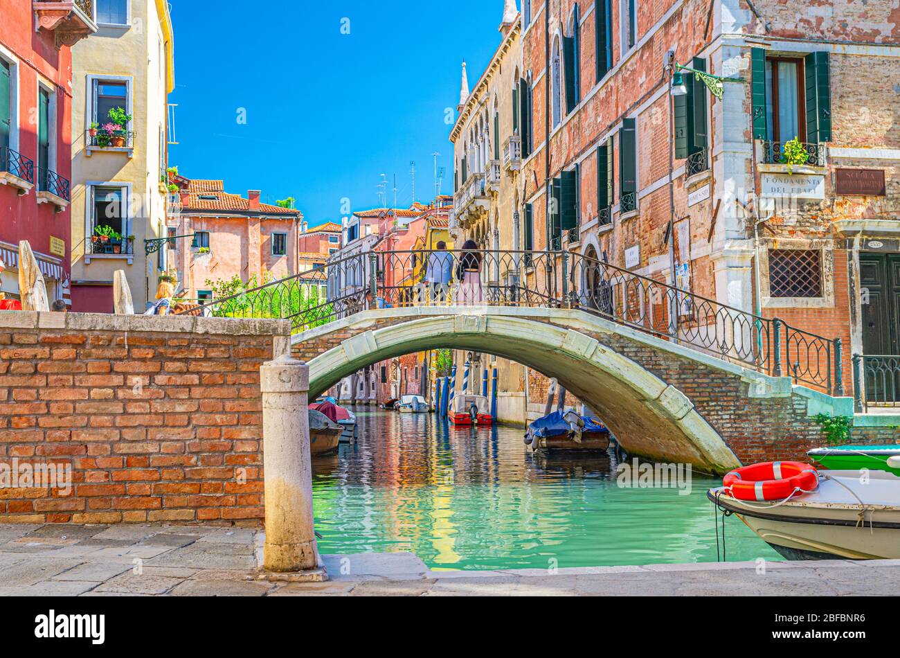 Ponte attraverso lo stretto canale d'acqua di Venezia con barche ormeggiate tra vecchi edifici colorati con balconi e pareti in mattoni, cielo blu, regione Veneto, N. Foto Stock