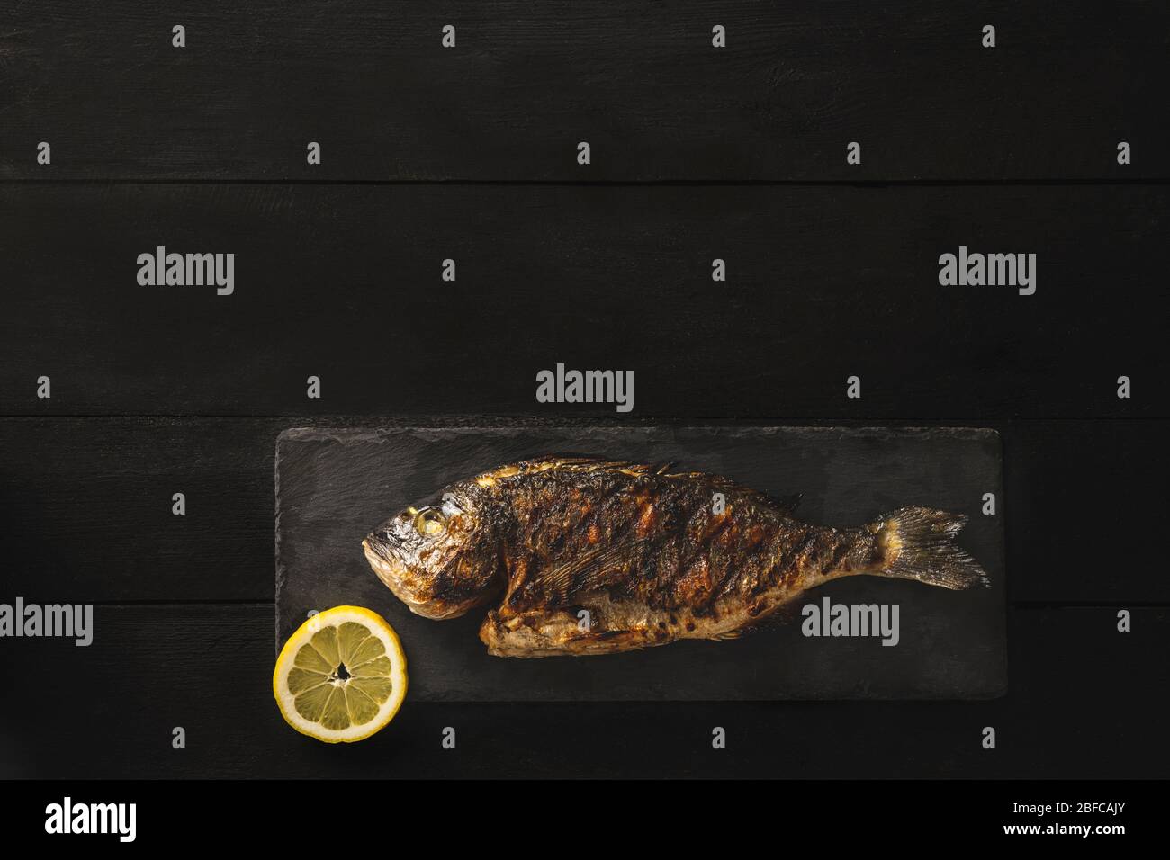 Pesce fresco o orata cotto su pietra nera con una fetta di limone su sfondo di legno nero Foto Stock