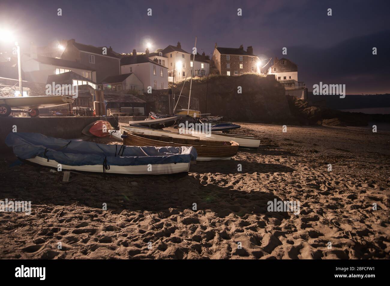 Barche tirate su sabbia per la notte con luci della strada circostanti che illuminano gli edifici e la spiaggia in una destinazione di vacanza nel Regno Unito di Cawsand. Foto Stock