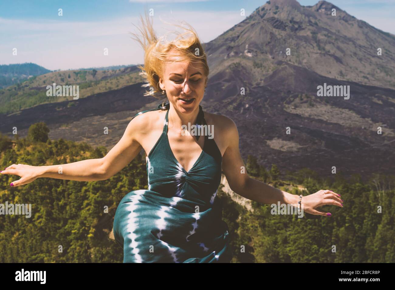 Giovane donna bionda con abito verde spalmato le mani e prendere il vento di fronte al vulcano Monte Batur sull'isola di Bali Foto Stock