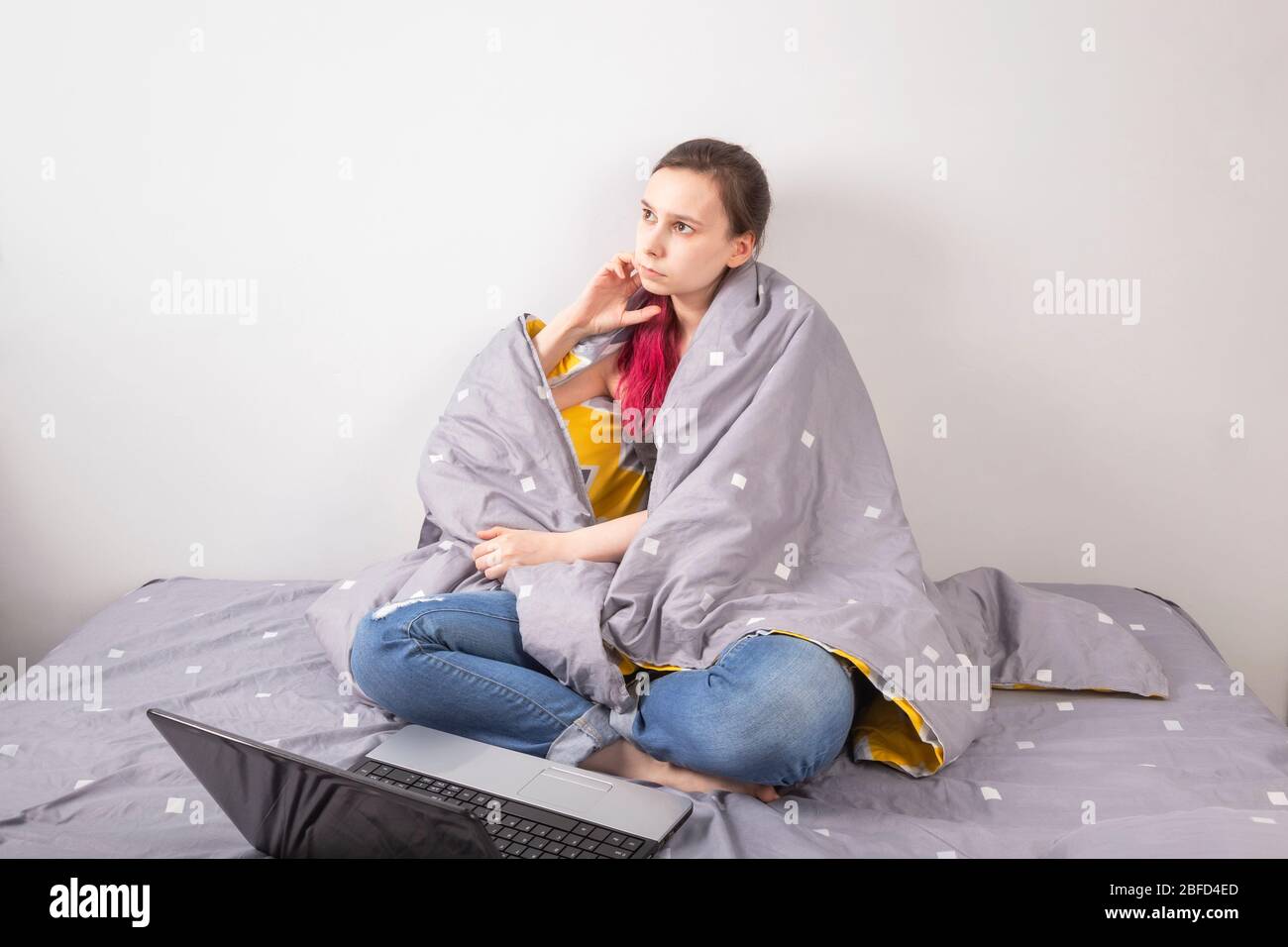 Concetto - lavoro da casa, autoisolamento, quarantena. La ragazza giovane siede nel letto coperto di coperta, guarda pensivamente lontano Foto Stock