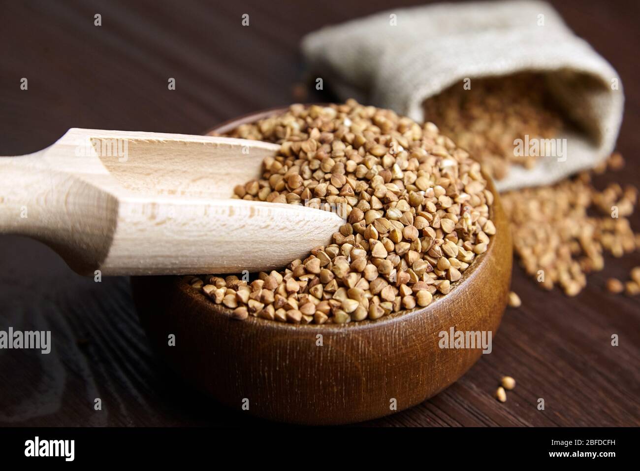 Semole di grano saraceno (semi di colmi) in ciotola di legno con cucchiaio, primo piano. Grano saraceno intero su sfondo legno, fuoco selettivo Foto Stock
