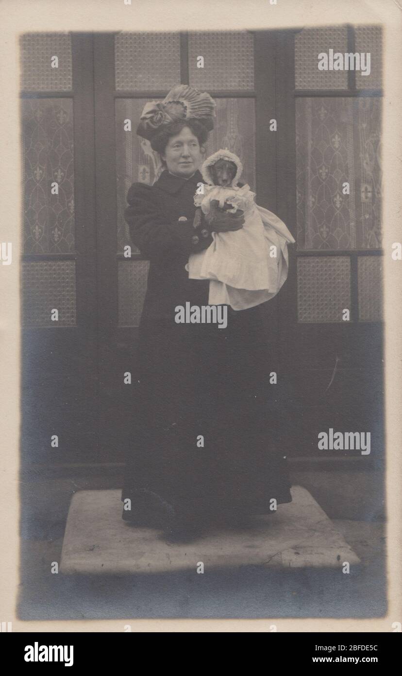 Vintage Edwardian 1910 cartolina fotografica di una signora vestito con intelligenza che tiene un piccolo cane nelle sue braccia. Il cane indossa un abito e un cofano Foto Stock