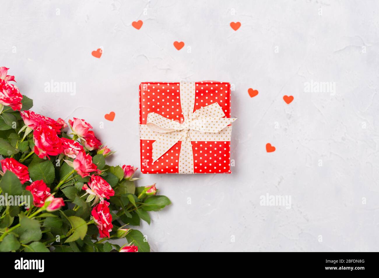 Confezione regalo rossa e rose rosse su sfondo bianco con confetti a cuore rosso. Regalo di Natale, sorpresa di San Valentino, idea di compleanno di festa della mamma. Piatto Foto Stock