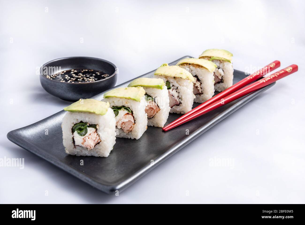 Rotolo di sushi con wrap avocado, salmone alla griglia, formaggio philadelphia e cipolle verdi contenitore nero e bacchette rosse. Foto Stock