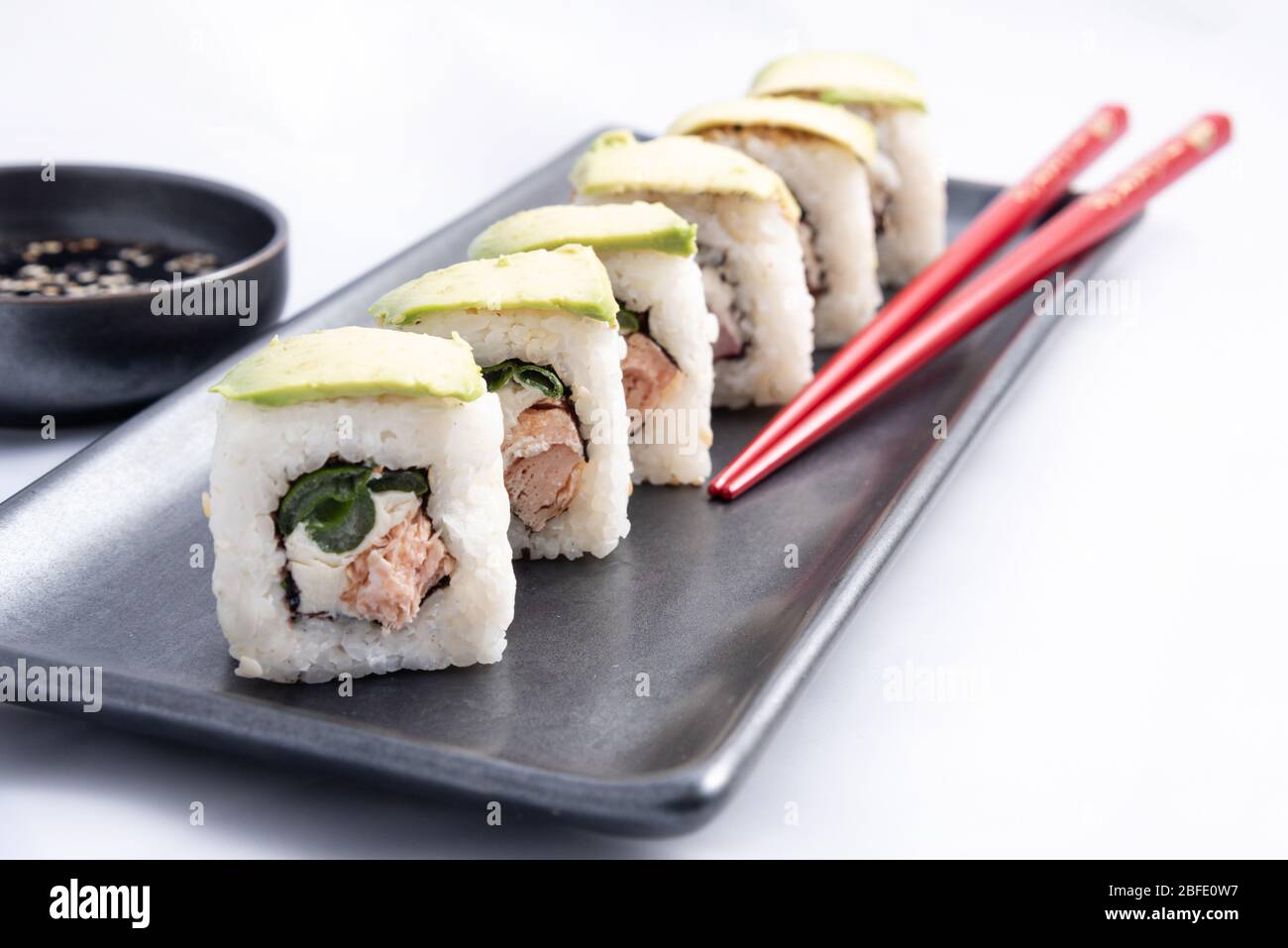 Rotolo di sushi con wrap avocado, salmone alla griglia, formaggio philadelphia e cipolle verdi contenitore nero e bacchette rosse. Foto Stock