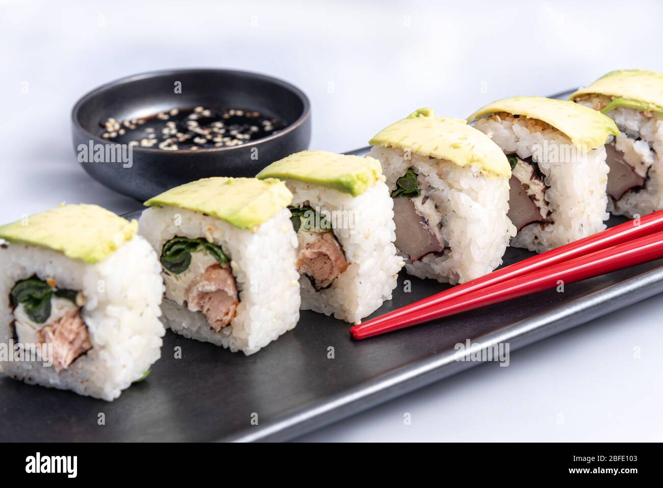 Rotolo di sushi con wrap avocado, salmone alla griglia, formaggio philadelphia e cipolle verdi contenitore nero e bacchette rosse. Foto Stock