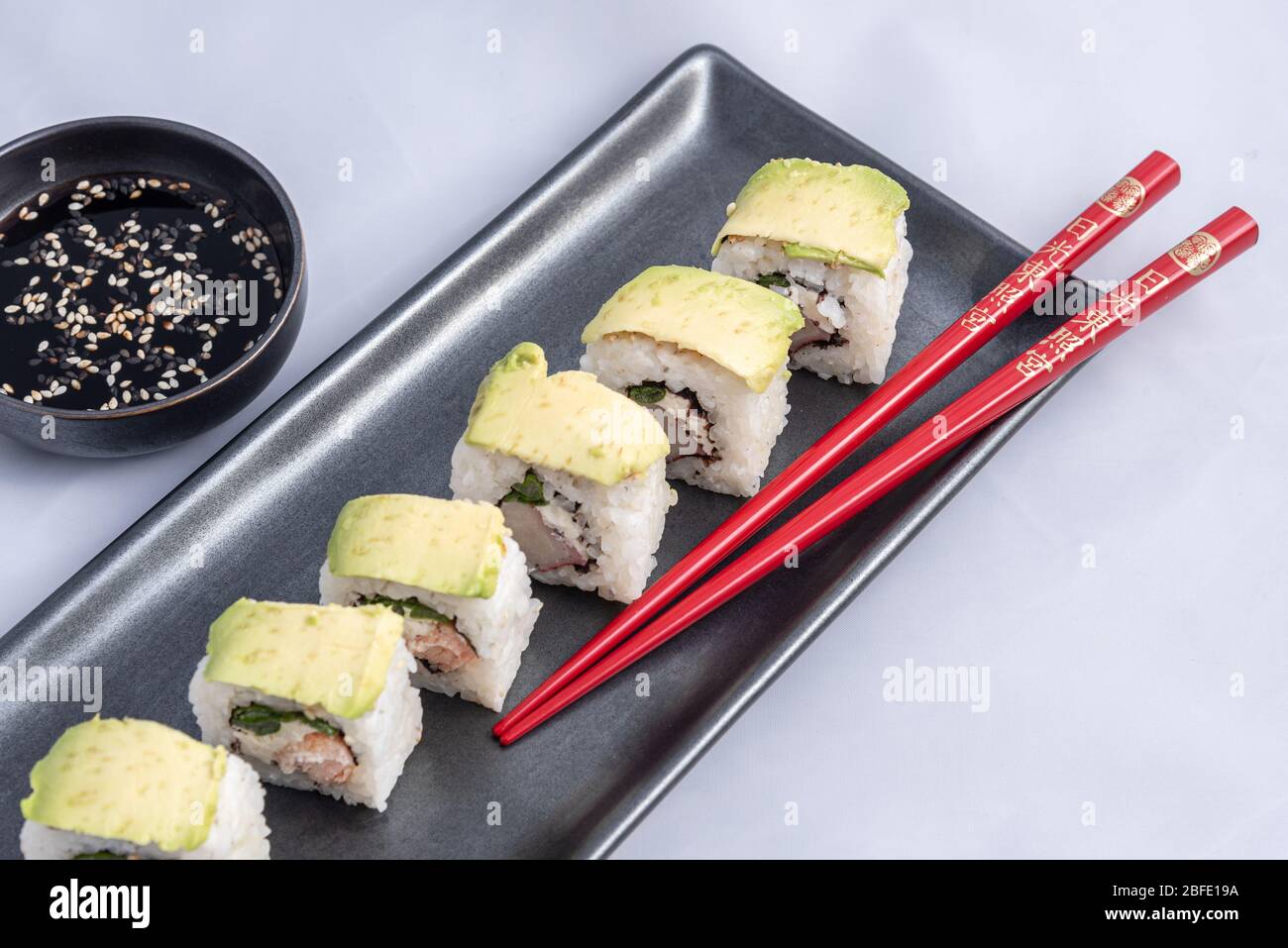 Rotolo di sushi con wrap avocado, salmone alla griglia, formaggio philadelphia e cipolle verdi contenitore nero e bacchette rosse. Foto Stock