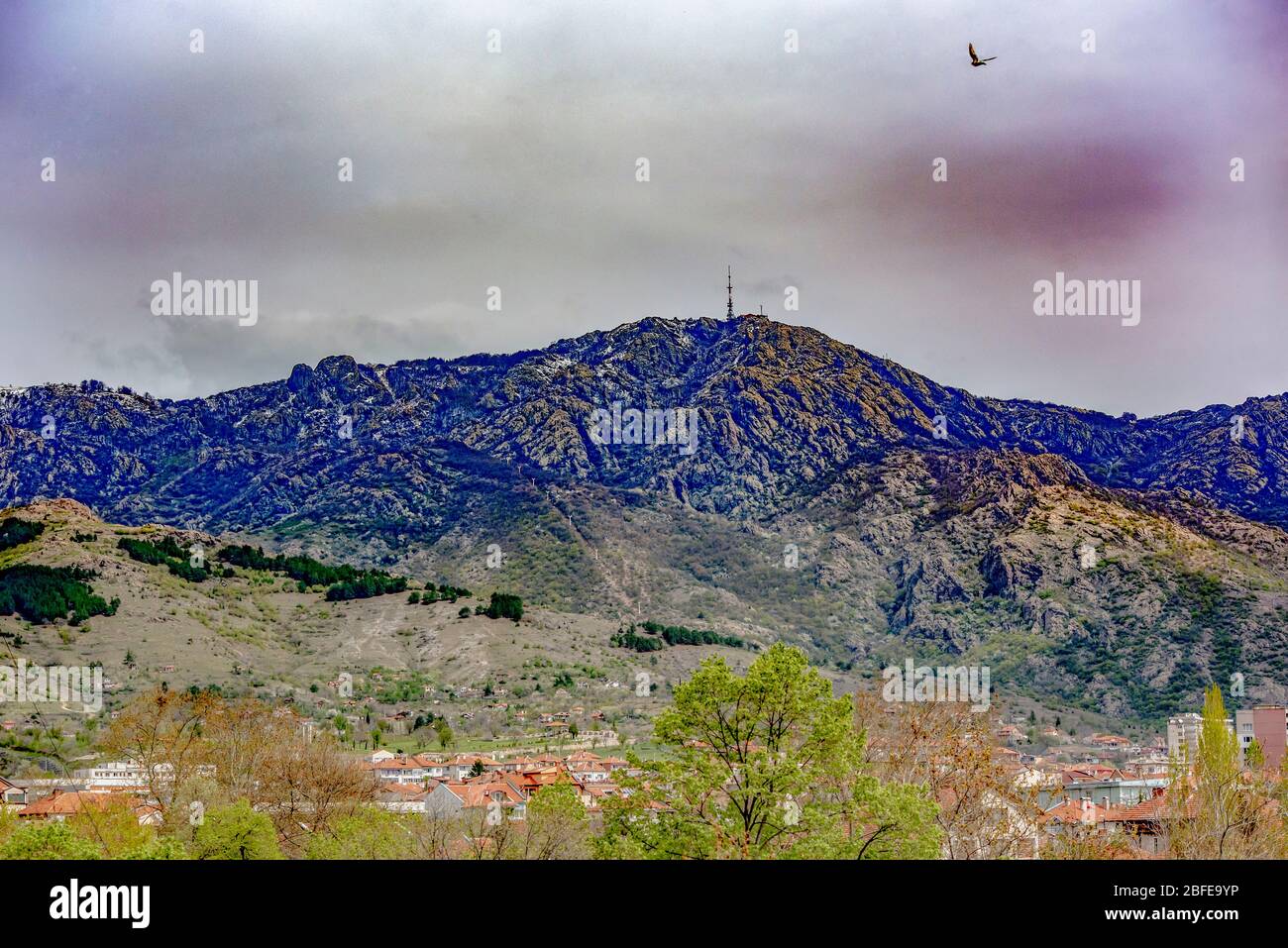 Vista spettacolare di una montagna rocciosa, cielo minaccioso e un uccello in volo. Città visibile ai piedi della montagna. Foto Stock