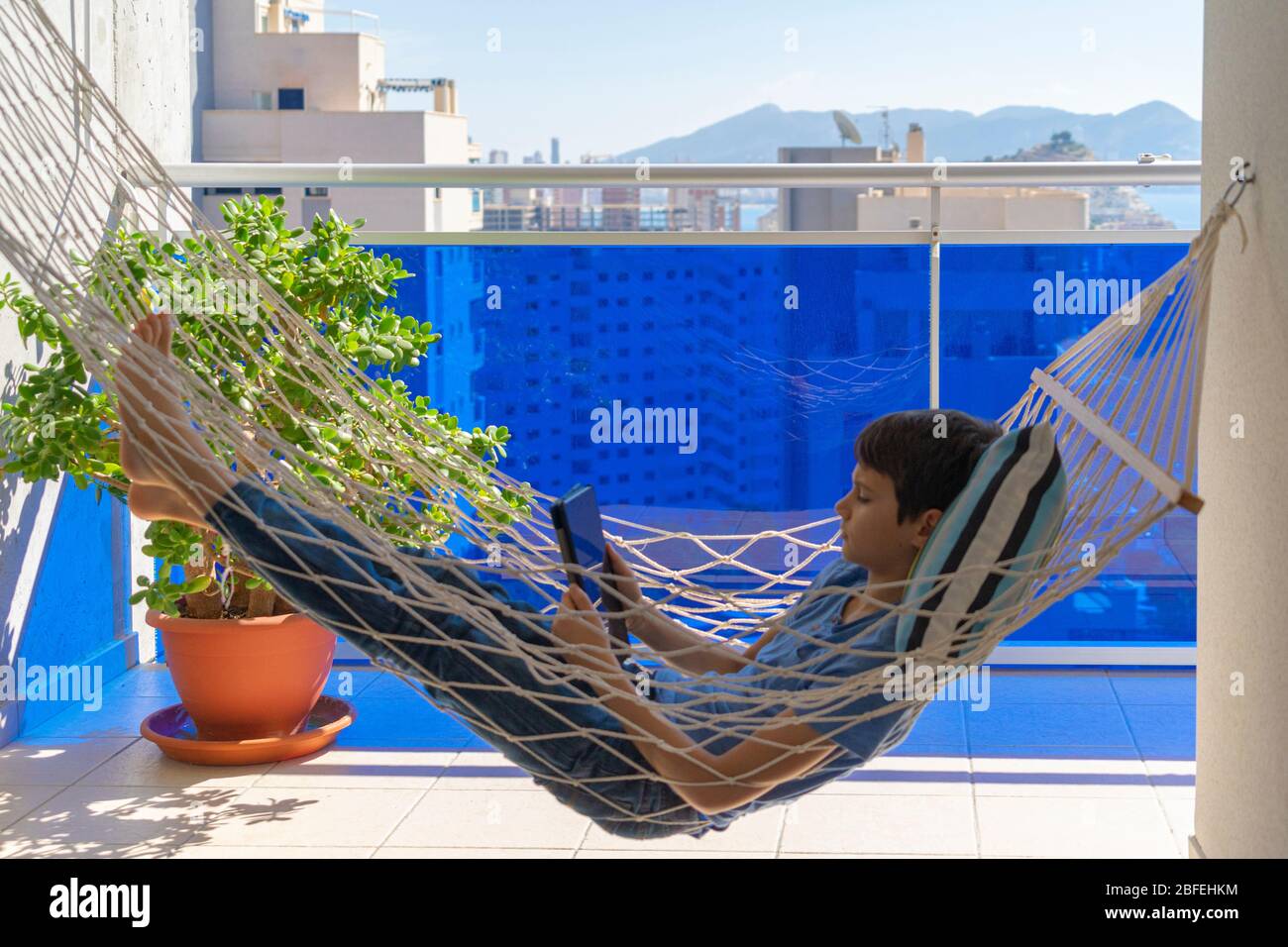 Ragazzo con computer tablet digitale che giace nell'amaca appesa sul balcone. Educazione, apprendimento a distanza online, homeschooling per i bambini Foto Stock