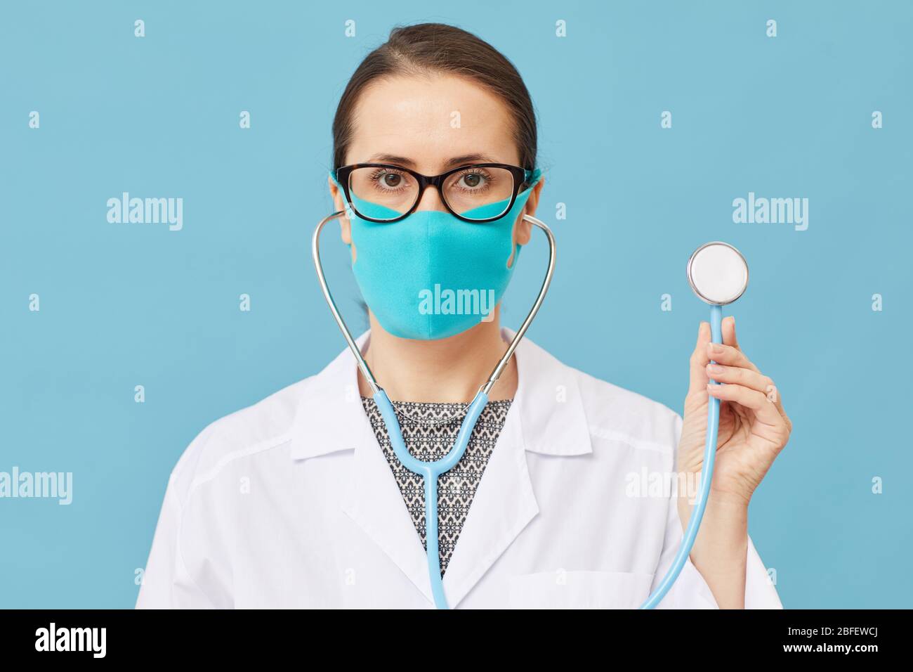 Ritratto di medico femminile in maschera protettiva che tiene stetoscopio e guardando la fotocamera su sfondo blu Foto Stock