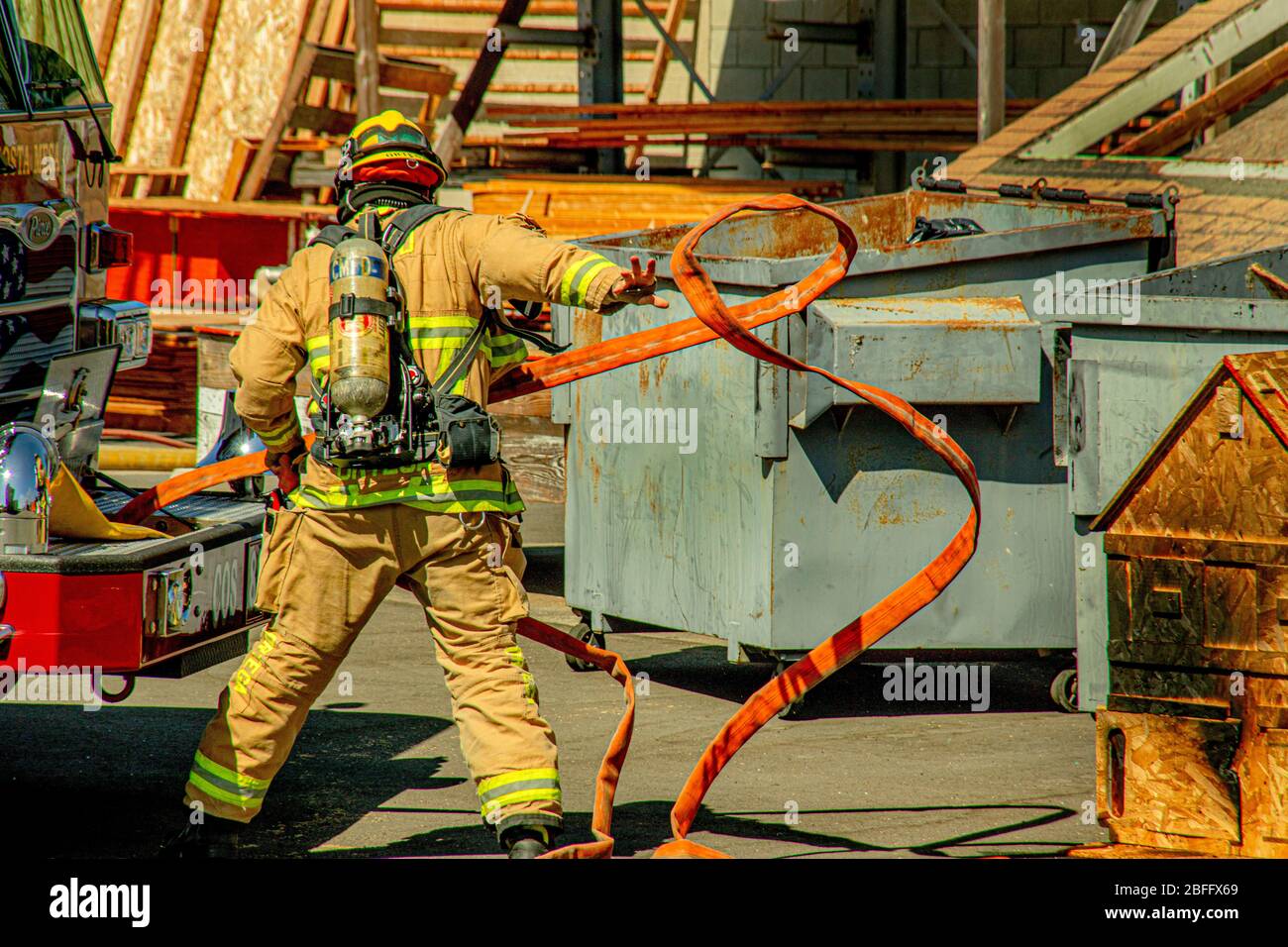 Con serbatoio d'aria e casco, un vigile del fuoco ispanico sarrotola rapidamente il tubo dell'acqua sulla scena di un blaze industriale a Costa Mesa, CA. Foto Stock