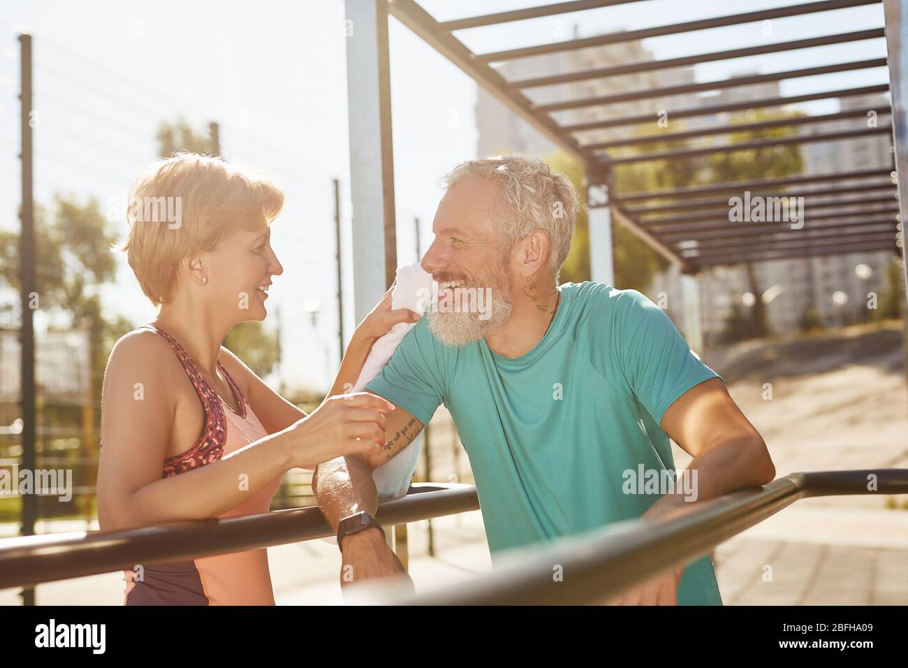 Riposo dopo l'allenamento. Donna sorridente che pulì il sudore del marito dopo aver fatto esercizio allo stadio. Senior Happy Family che lavora insieme nel Foto Stock