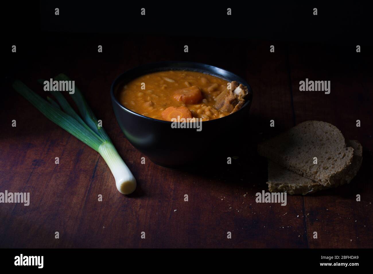 Costolette di maiale affumicate con fagioli e pezzi di carota, in una ciotola nera, con due pezzi di pane a grani interi a fianco, e una cipolla verde Foto Stock