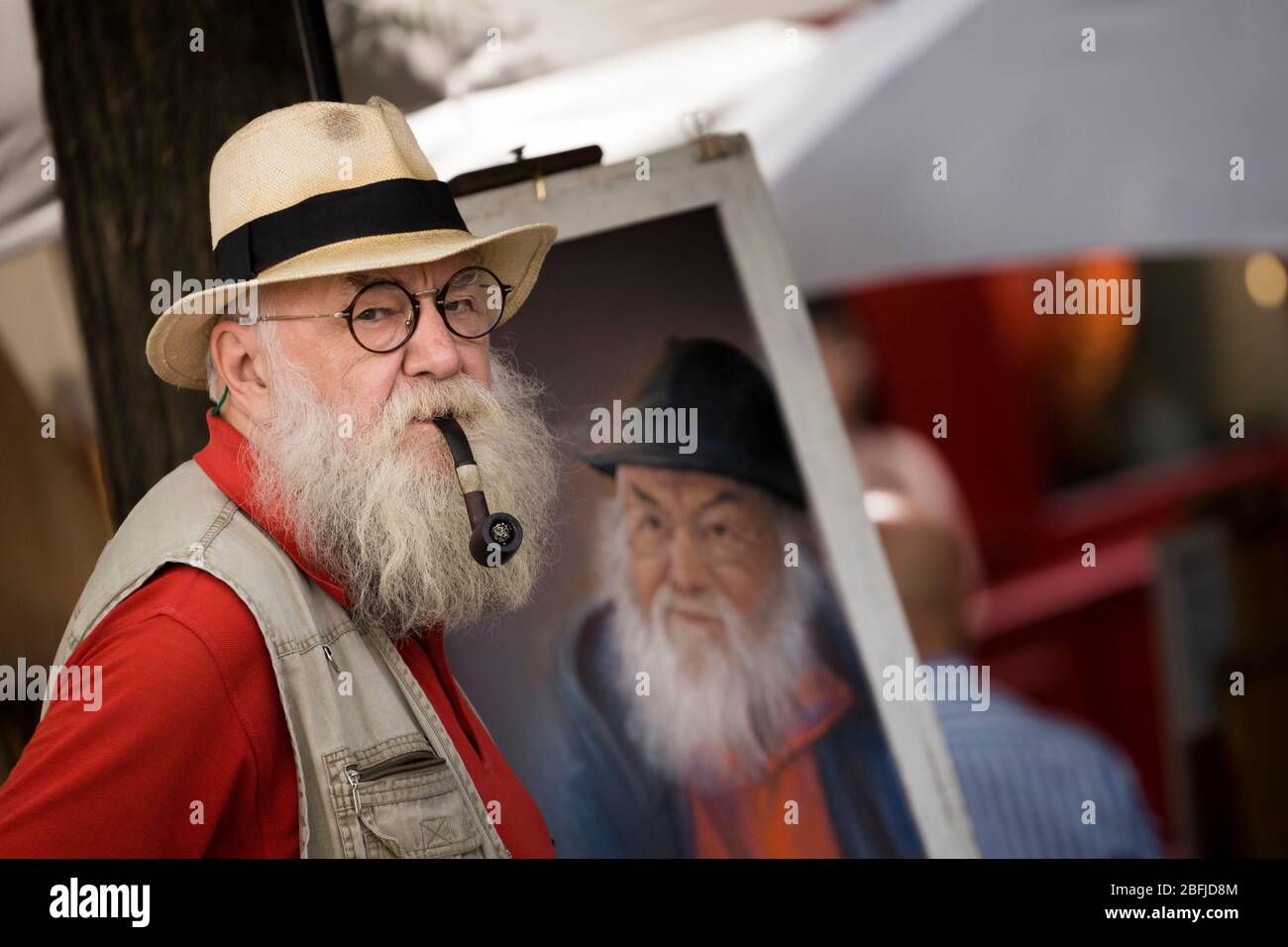 Ritratto di un artista anziano maschile con un cappello, una pipa fumante e una barba, davanti al suo cavalletto al mercato degli artisti di Montmartre, Parigi Foto Stock