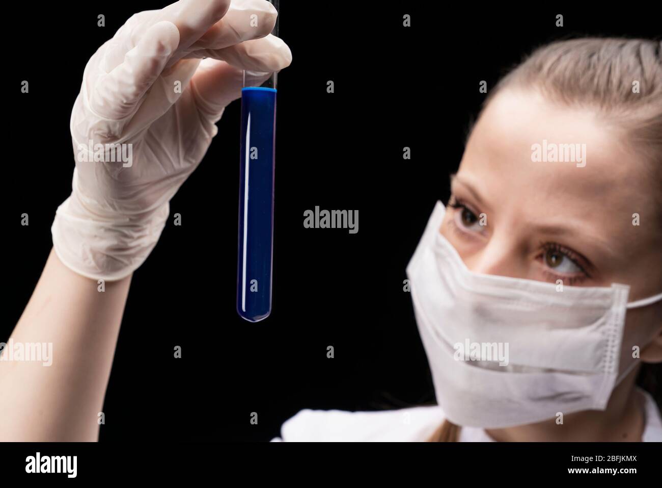 una provetta con una sostanza luminosa blu liquida nelle mani di una giovane donna in una maschera, uno specialista scientifico o studente laureato Foto Stock