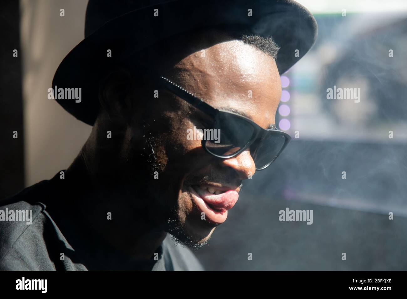 Ragazzo nero con occhiali da sole e cappello vintage circondato da fumo  mentre sorride e tocca il labbro con la lingua Foto stock - Alamy