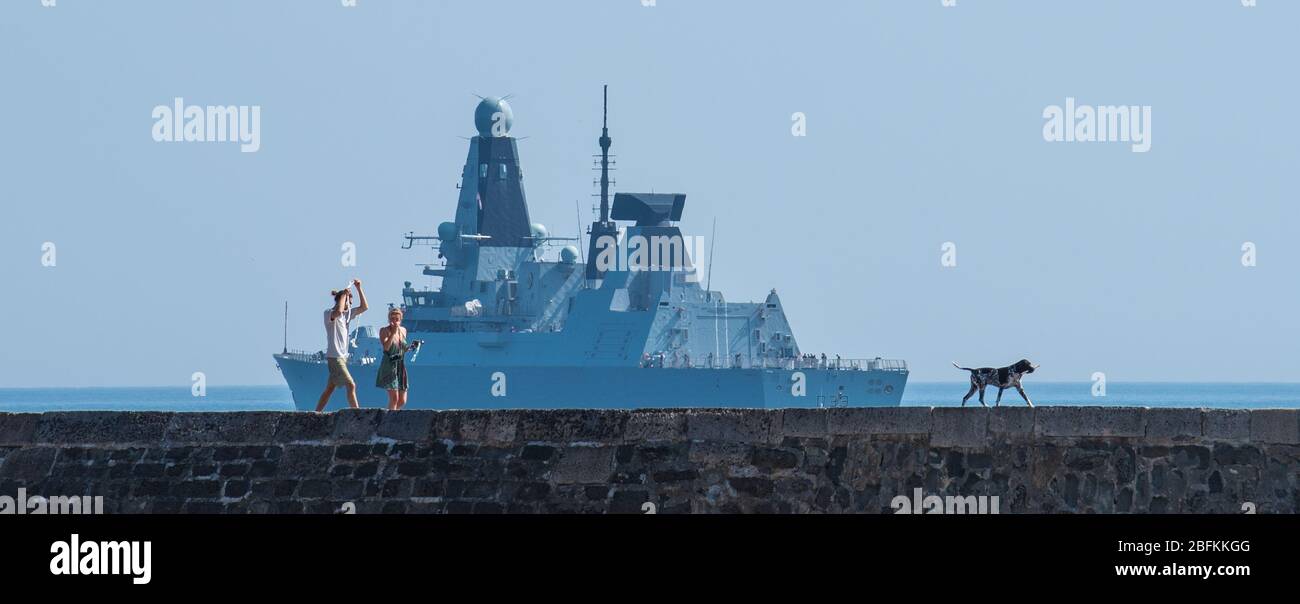 Lyme Regis, Dorset, Regno Unito. 19 Apr 2020. Tempo in Gran Bretagna: Royal Navy nave da guerra HMS Dauntless ancorato a Lyme Regis in un pomeriggio caldo e soleggiato durante il blocco del coronavirus. Una coppia cammina lungo il muro di Cobb con il loro cane. Credit: Celia McMahon/Alamy Live News Foto Stock