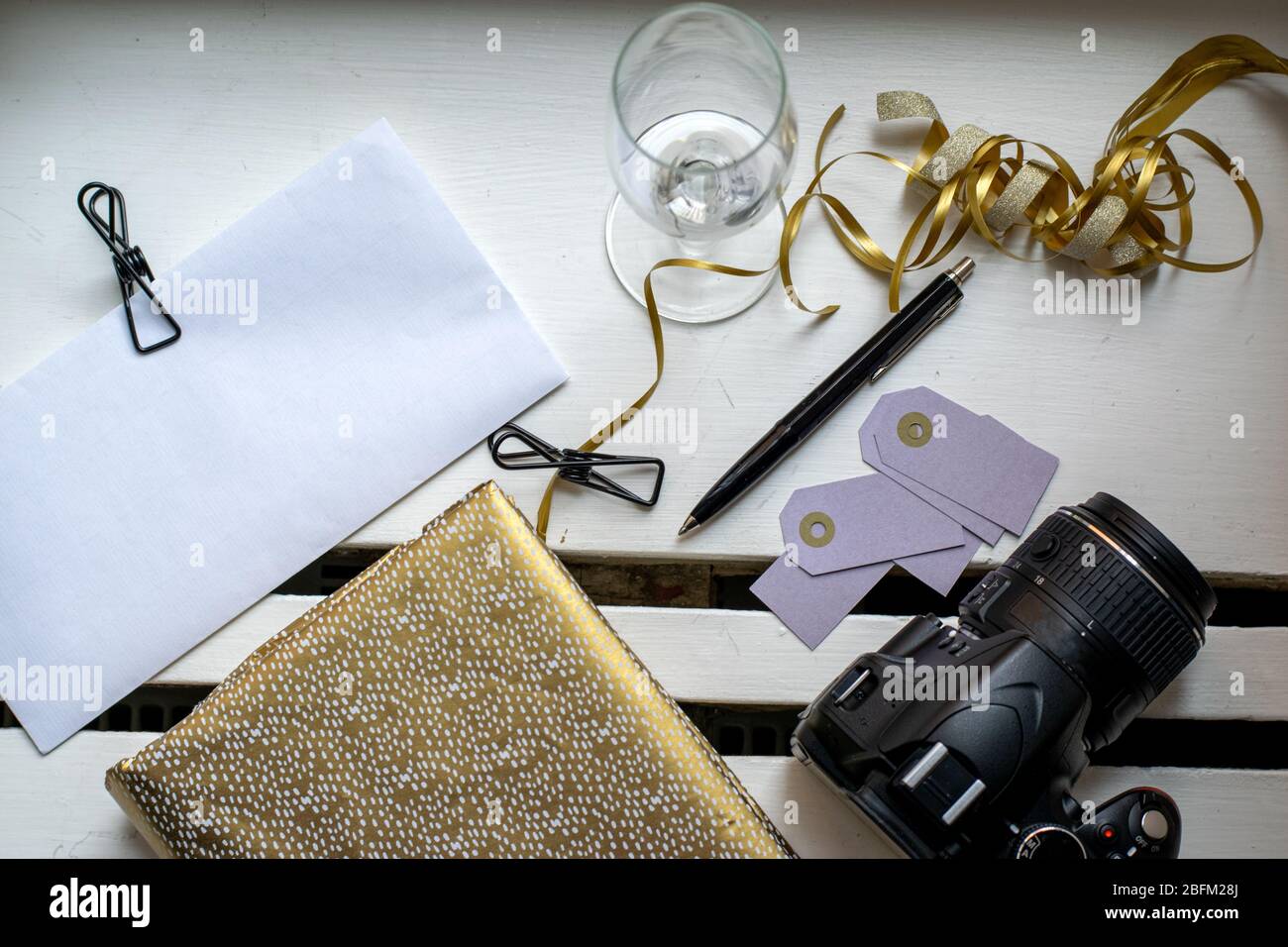 Preparazione per le feste con notebook dorato, bicchiere di champagne vuoto con penna, fotocamera e clip per rilegatura su busta bianca Foto Stock