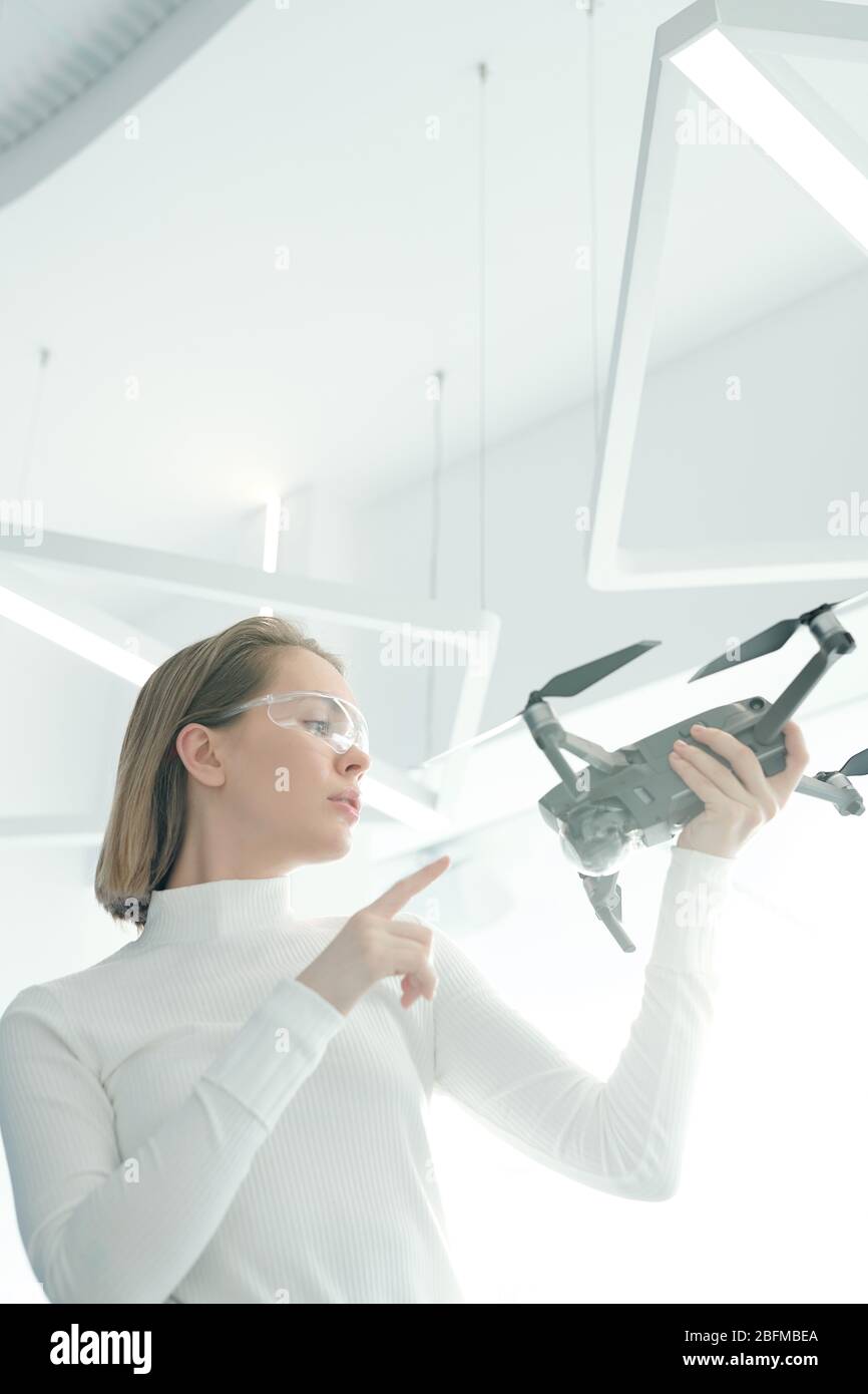 Giovane donna in occhiali intelligenti che tiene il drone davanti a sé mentre impara come inizia e funziona in laboratorio futuristico Foto Stock