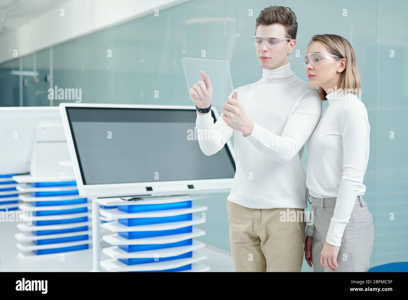 Giovane uomo intelligente e sua collega con occhiali intelligenti che guardano il display di un tablet trasparente mentre scansionano nuove informazioni Foto Stock
