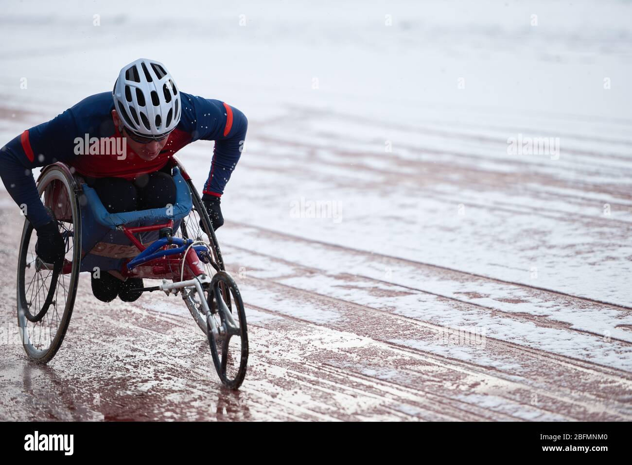 Corse su sedia a rotelle. Atleta maschile fisicamente non vedente in addestramento su casco in carrozzina da corsa su pista coperta di neve sciogliente in condizioni atmosferiche avverse Foto Stock