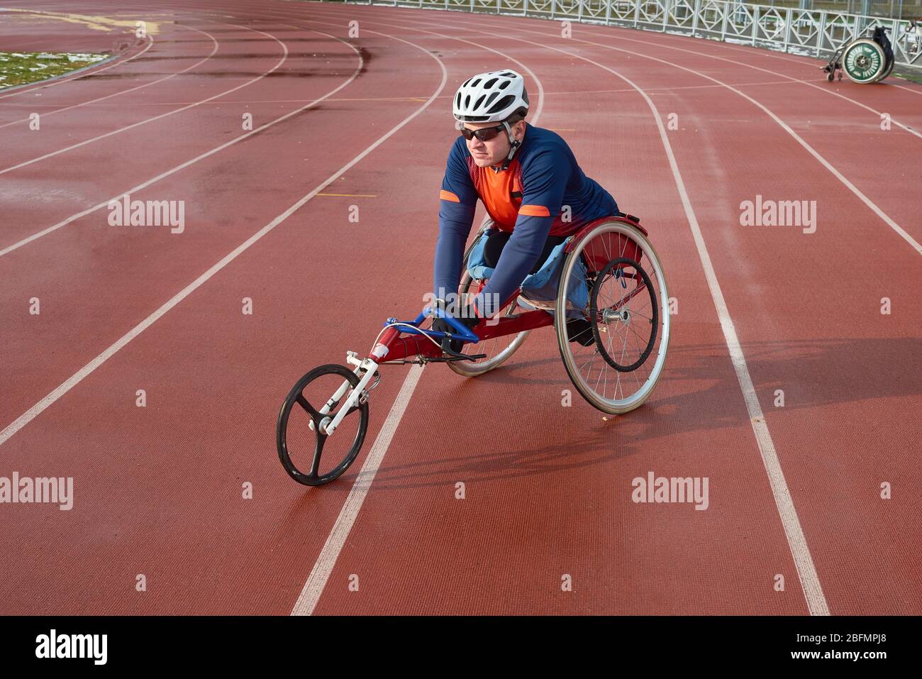 Sportivo con disabilità in casco e occhiali da sole seduto in sedia a rotelle da corsa al campo e pista all'aperto e pronto per la maratona Foto Stock